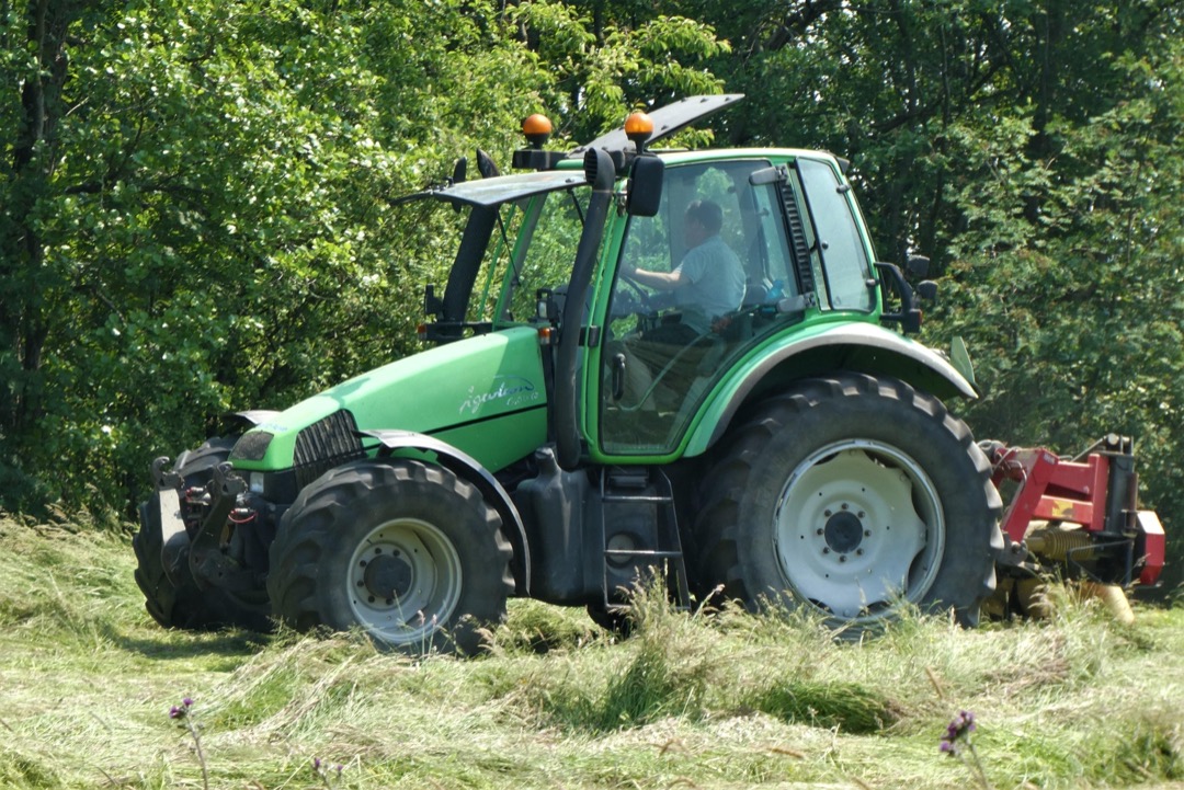 Foto van een maaiende grasmaaier-tractor op een grasveld voor een bomenrij.